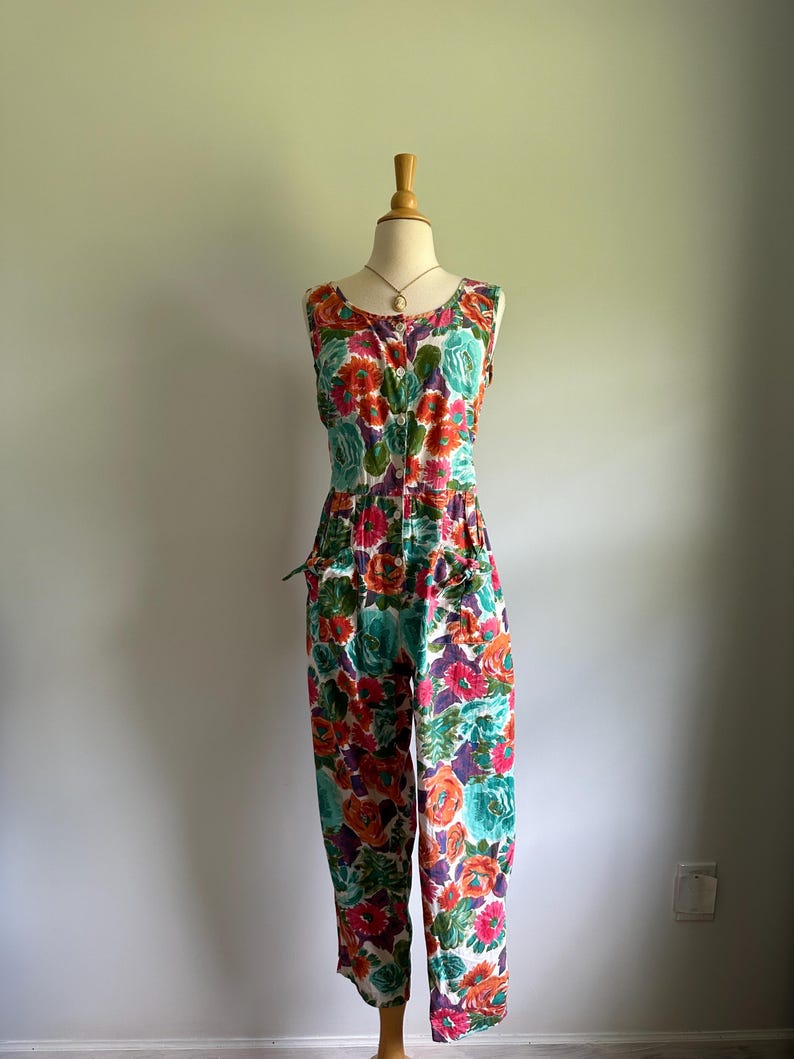 May include: A sleeveless floral jumpsuit in a vibrant pattern of red, orange, teal, and purple flowers on a white background. The jumpsuit features a button-down front, pockets, and a fitted waist. The garment is displayed on a mannequin.