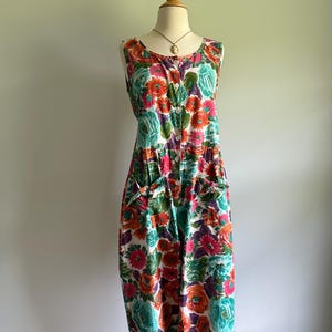 May include: A sleeveless floral jumpsuit in a vibrant pattern of orange, pink, teal, and purple flowers on a white background. The jumpsuit features a button-down front, pockets, and is displayed on a mannequin.