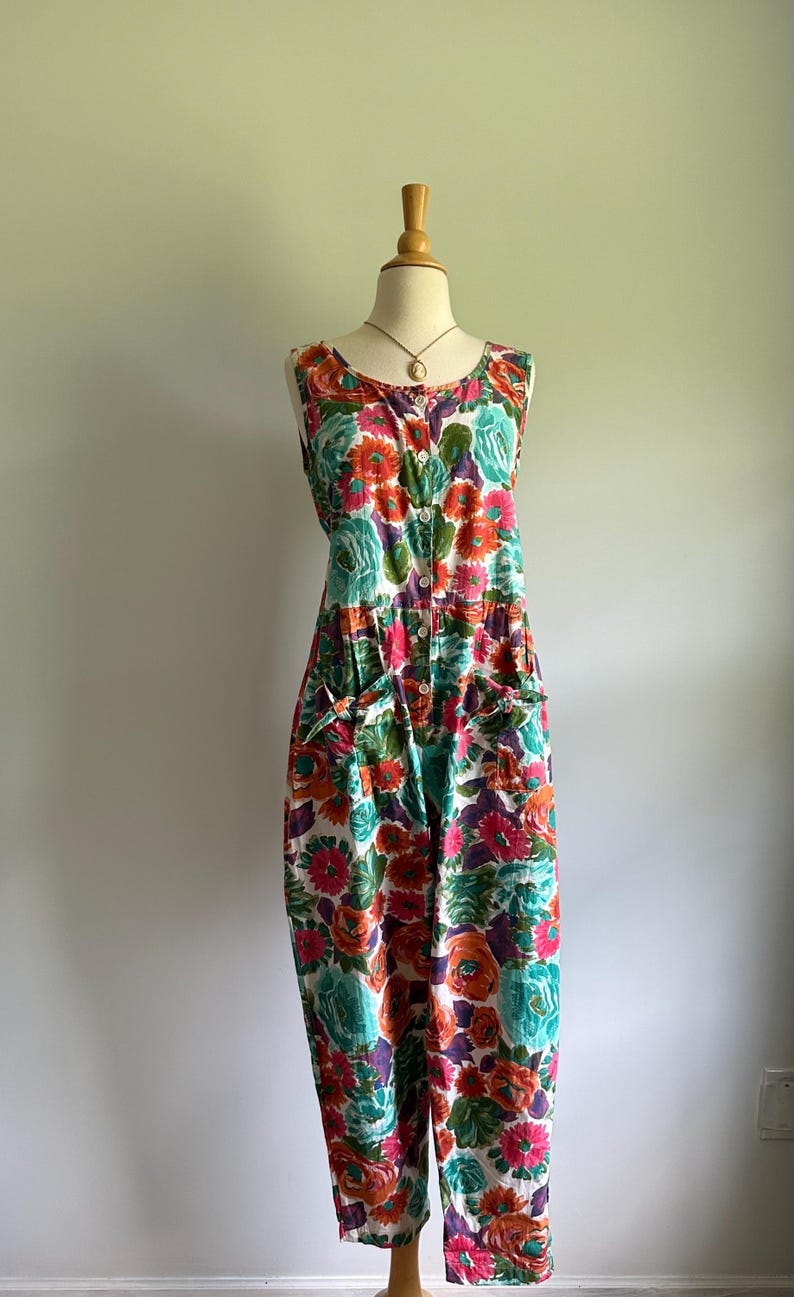 May include: A sleeveless floral jumpsuit in a vibrant pattern of orange, pink, teal, and purple flowers on a white background. The jumpsuit features a button-down front, pockets, and is displayed on a mannequin.