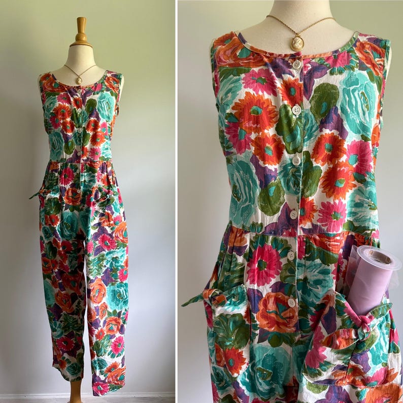 May include: A sleeveless jumpsuit with a vibrant floral pattern in shades of pink, orange, teal, and purple. The jumpsuit features a button-down front, pockets, and a round neckline. The garment is displayed on a mannequin.