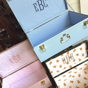 May include: Three metal storage trunks in pastel colors: light blue, pink, and olive green. Each trunk has a gold handle and clasp. The interiors are lined with patterned fabric, including gingham, floral, and teddy bear prints. Monogrammed initials are embroidered on the inside lids.