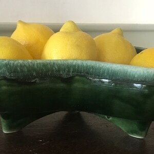May include: A green ceramic bowl with four lemons inside. The bowl has a white glaze and three legs.