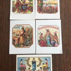 May include: Four vintage cigar labels featuring illustrations of Native American figures and text. The labels are printed on white paper with a yellowed tint. The labels are titled "Alpha", "Americanos", "Tropical Goddess", and "Susquehanna". The fifth label features a woman in a blue gown standing on a boat with two cherubs rowing. The text on the label reads "Tabacos Muy Superiores".