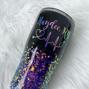 May include: Black tumbler with a rainbow glitter design and the text "Haylee, RN" with a stethoscope and heartbeat graphic.