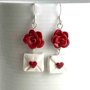 May include: A pair of dangle earrings featuring a red rose and a white envelope with a red heart. The earrings are made of polymer clay and have silver hooks.