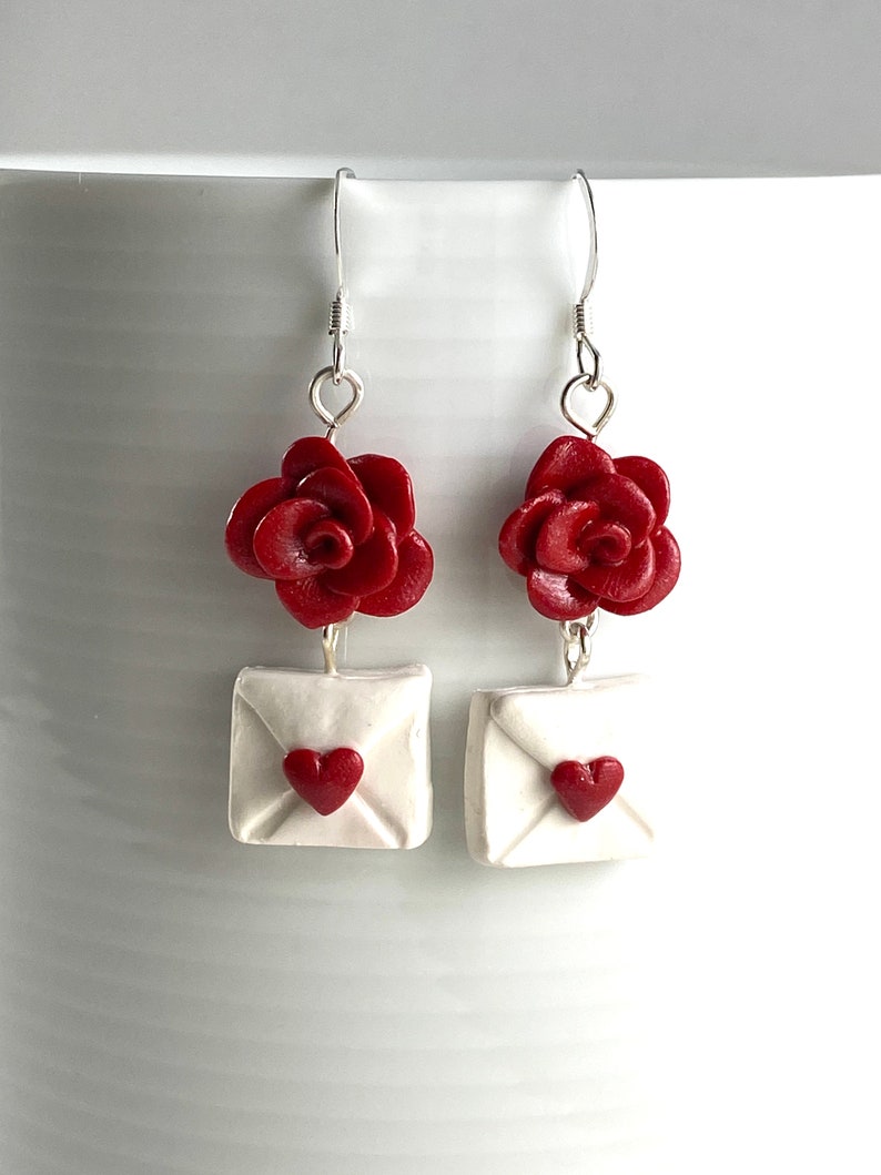 May include: A pair of dangle earrings featuring a red rose and a white envelope with a red heart. The earrings are made of polymer clay and have silver hooks.