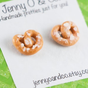 May include: A pair of salt-covered pretzel earrings. The earrings are made of polymer clay and are a light brown color. The earrings are on a white card with the text "jennyoandco.etsy.com" printed on it.