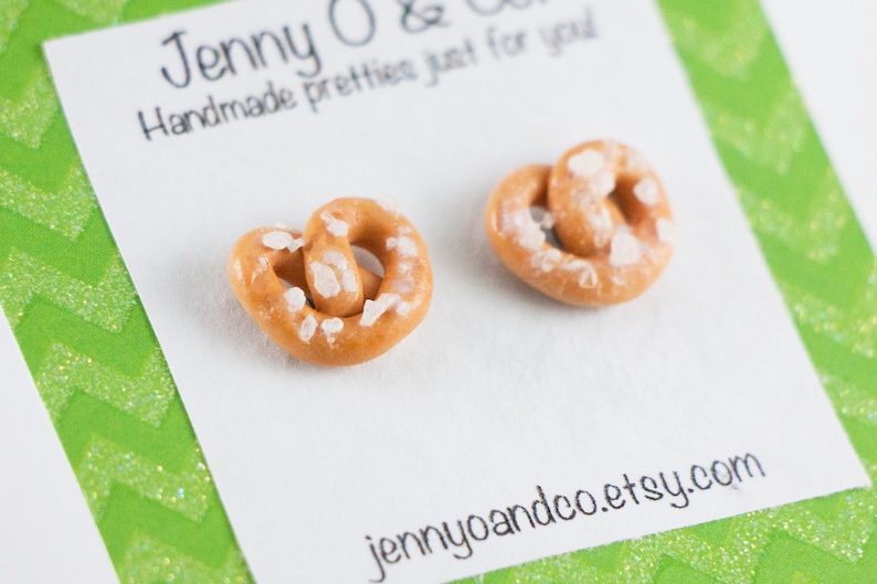 May include: A pair of salt-covered pretzel earrings. The earrings are made of polymer clay and are a light brown color. The earrings are on a white card with the text "jennyoandco.etsy.com" printed on it.