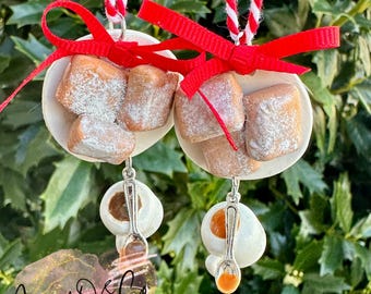 May include: Two handmade Christmas ornaments. Each ornament features a white shell-shaped base with brown, powdered sugar-dusted treats and a red bow. Below, a white egg-shaped charm with a silver spoon hangs. The text "Jenny O & Co" is visible.