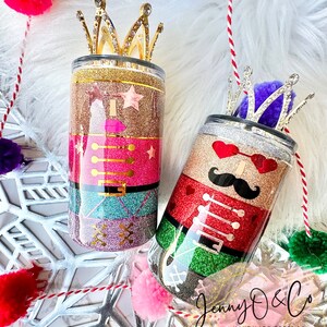 May include: Two glitter-covered tumblers with a nutcracker theme. One tumbler is pink, gold, and green with a gold crown on top. The other tumbler is red, green, and silver with a silver crown on top. The tumblers are decorated with glitter and have a nutcracker design. The text "Jenny O & Co. Handmade Pretties. Just for You." is visible at the bottom of the image.