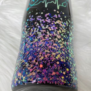 May include: Black tumbler with a teal and white design that says "Mydee, RN" with a heart and a heartbeat graphic. The tumbler is covered in iridescent glitter.