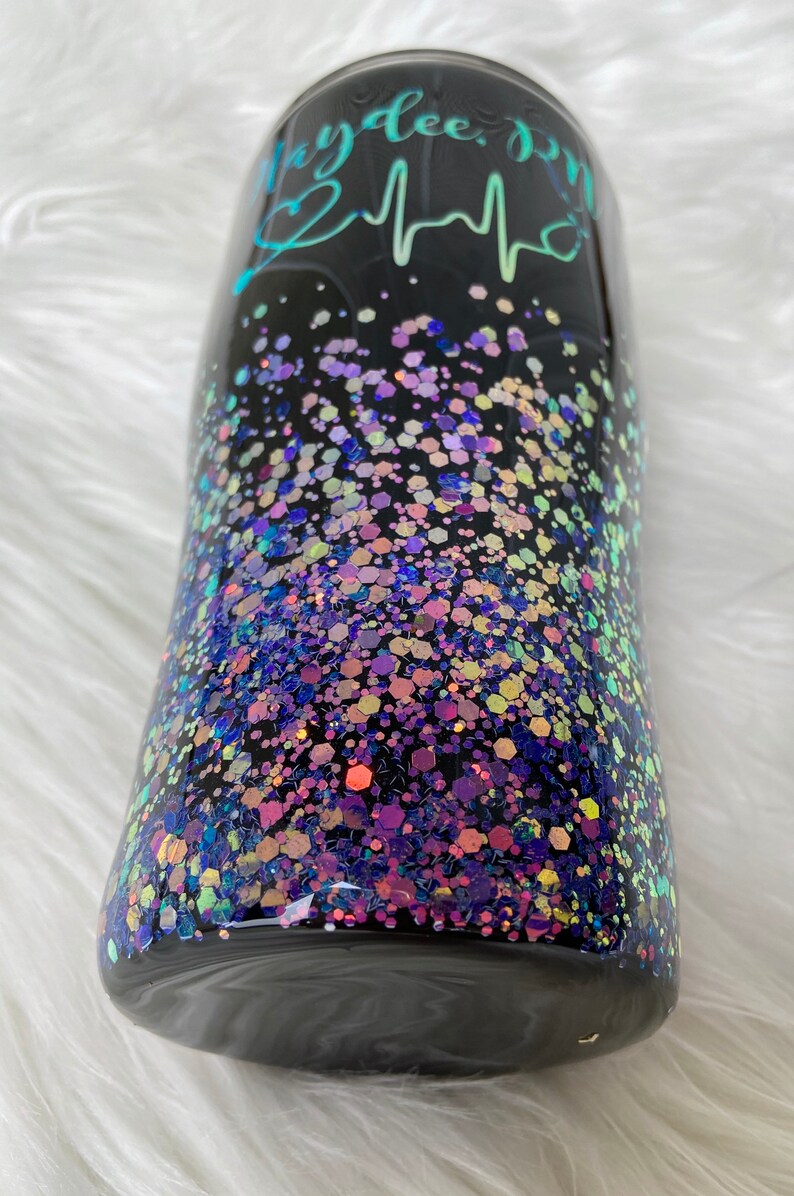 May include: Black tumbler with a teal and white design that says "Mydee, RN" with a heart and a heartbeat graphic. The tumbler is covered in iridescent glitter.