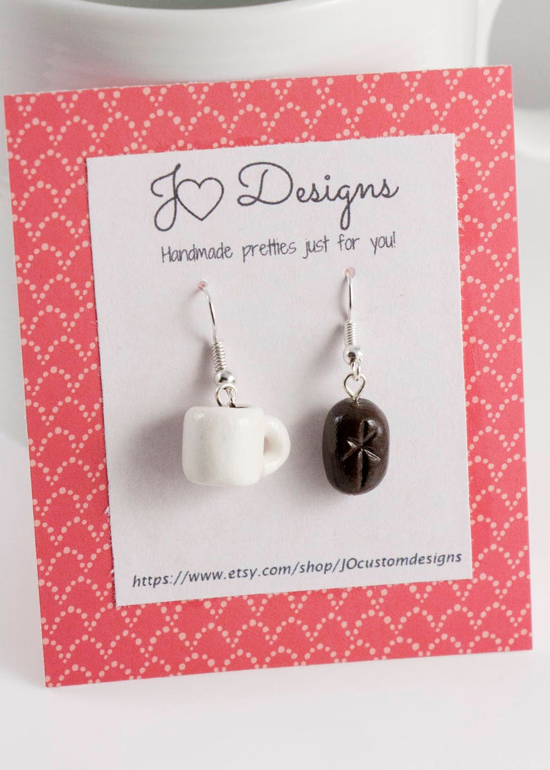 May include: A pair of dangle earrings featuring a white coffee mug and a brown coffee bean charm. The earrings are displayed on a pink and white card with the text "J &hearts; Designs Handmade pretties just for you!" and a website address.