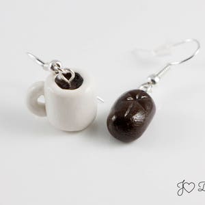 May include: A pair of earrings featuring a white ceramic coffee cup with brown coffee inside and a brown coffee bean. The earrings are silver and have a hook closure.
