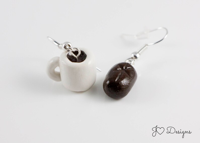 May include: A pair of earrings featuring a white ceramic coffee cup with brown coffee inside and a brown coffee bean. The earrings are silver and have a hook closure.