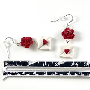 May include: A pair of earrings featuring a red rose and a white envelope with a red heart. The earrings are approximately 1 1/2 inches long.