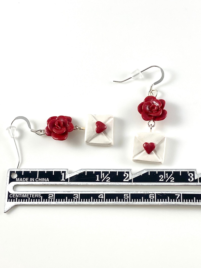 May include: A pair of earrings featuring a red rose and a white envelope with a red heart. The earrings are approximately 1 1/2 inches long.