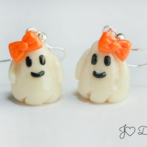 May include: A pair of white ghost earrings with orange bows. The ghosts have black eyes and a black smile.