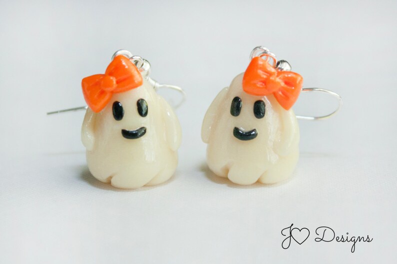 May include: A pair of white ghost earrings with orange bows. The ghosts have black eyes and a black smile.