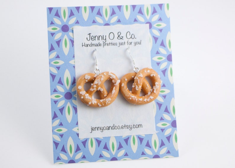 Puede incluir: Un par de pendientes de plata con forma de pretzel con un dije de pretzel cubierto de sal. Los pendientes se muestran en una tarjeta con estampado azul y blanco con el texto "Jenny O & Co. Handmade pretties just for you! jennyoandco.etsy.com".