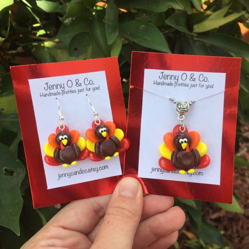 May include: A set of Thanksgiving-themed jewelry featuring a pair of earrings and a necklace. Both pieces feature a brown turkey with red, orange, and yellow feathers. The earrings are silver and have a hook closure. The necklace has a silver chain and a silver clasp. The cards are red and have the text "Jenny O & Co. Handmade pretties just for you!" and the website address "jennyoandeo.etsy.com".