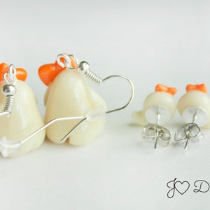 May include: A pair of dangle earrings featuring white ceramic chicks with orange beaks and combs. The earrings are on a white background.