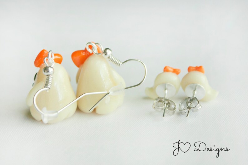 May include: A pair of dangle earrings featuring white ceramic chicks with orange beaks and combs. The earrings are on a white background.