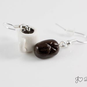 May include: A pair of earrings featuring a white ceramic coffee mug and a brown coffee bean charm. The earrings are silver and have a hook closure.
