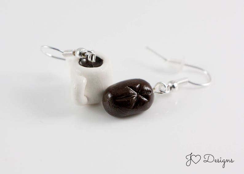May include: A pair of earrings featuring a white ceramic coffee mug and a brown coffee bean charm. The earrings are silver and have a hook closure.