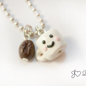 May include: A silver chain necklace with a white ceramic coffee cup charm with a smiling face and two pink cheeks. The charm is attached to a brown coffee bean charm.