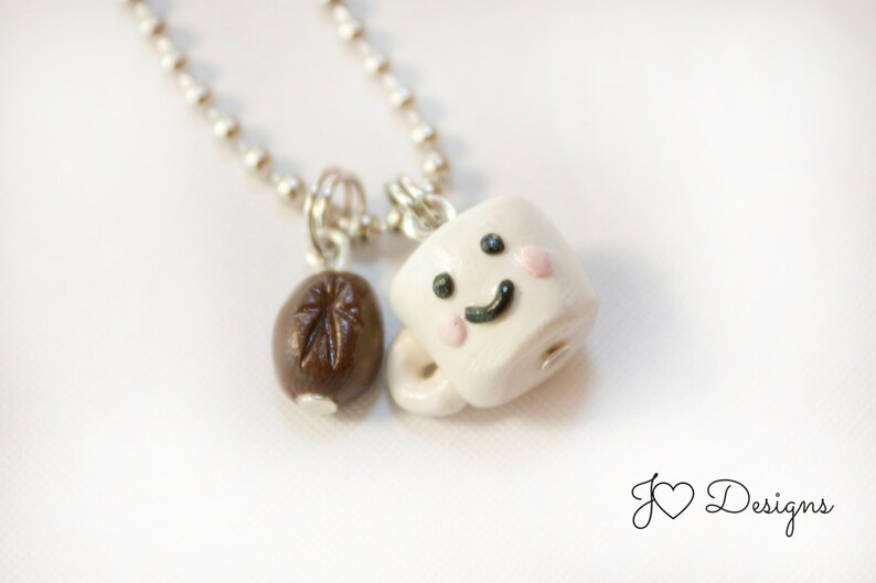 May include: A silver chain necklace with a white ceramic coffee cup charm with a smiling face and two pink cheeks. The charm is attached to a brown coffee bean charm.