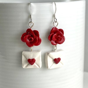 May include: Red rose flower dangle earrings with a white envelope charm featuring a red heart. The earrings are made of polymer clay.