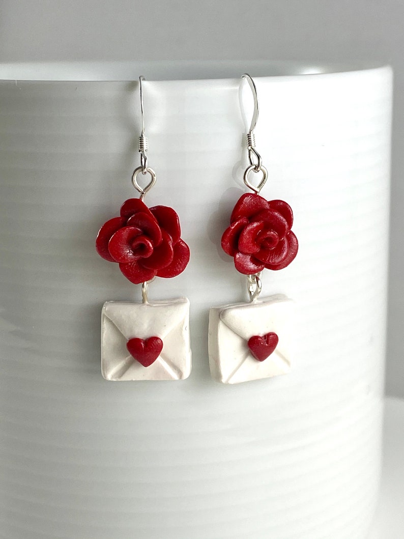 May include: Red rose flower dangle earrings with a white envelope charm featuring a red heart. The earrings are made of polymer clay.