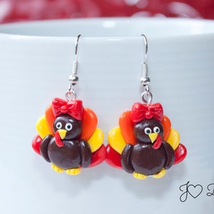 May include: A pair of dangle earrings featuring a brown turkey with a red bow, yellow and orange feathers, and silver hooks.