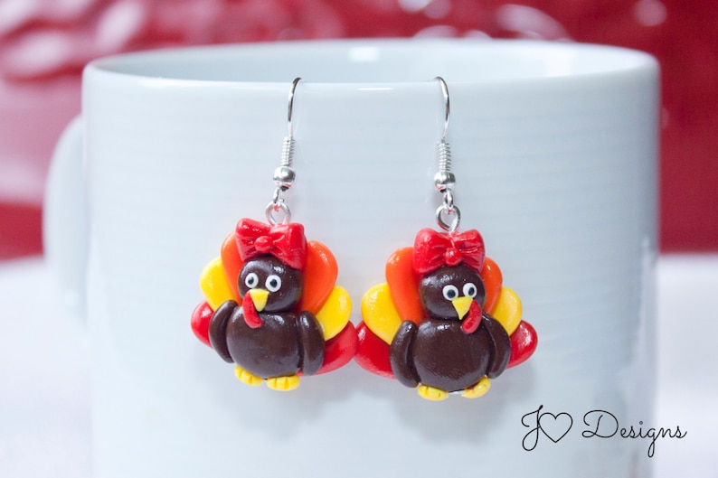 May include: A pair of dangle earrings featuring a brown turkey with a red bow, yellow and orange feathers, and silver hooks.