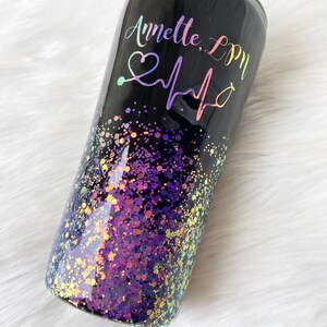 May include: Black tumbler with a purple and gold glitter design. The tumbler has the text "Annette, LPN" and a heart with a stethoscope and heartbeat graphic.