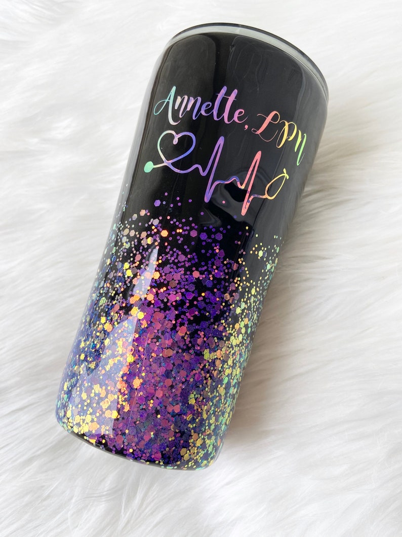 May include: Black tumbler with a purple and gold glitter design. The tumbler has the text "Annette, LPN" and a heart with a stethoscope and heartbeat graphic.