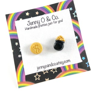May include: A pair of gold and black polymer clay earrings. One earring is a gold coin with a dollar sign on it. The other earring is a black pot filled with gold coins. The earrings are displayed on a card with a rainbow and star pattern. The card also has the text "Jenny O & Co. Handmade pretties just for you!" and the website address "jennyoandco.etsy.com".