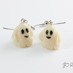 May include: A pair of white ghost earrings with black eyes and a smiling mouth. The earrings are made of polymer clay and have silver hooks.