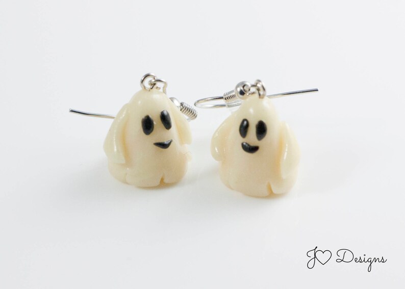 May include: A pair of white ghost earrings with black eyes and a smiling mouth. The earrings are made of polymer clay and have silver hooks.