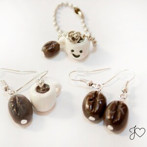May include: Two pairs of earrings featuring a white ceramic coffee cup charm and a brown coffee bean charm. The coffee cup charms have a happy face painted on them.