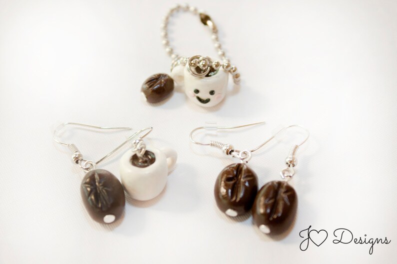 May include: Two pairs of earrings featuring a white ceramic coffee cup charm and a brown coffee bean charm. The coffee cup charms have a happy face painted on them.