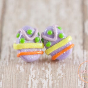 K&ouml;nnte beinhalten: Ein Paar violette Osterei-Ohrringe mit gelben, orangefarbenen und gr&uuml;nen Akzenten. Die Ohrringe sind aus Polymer Clay gefertigt und haben eine glitzernde Oberfl&auml;che.