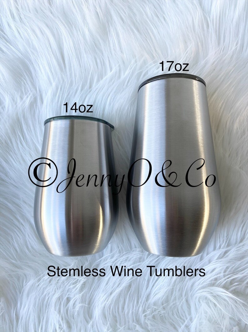 May include: Two stainless steel stemless wine tumblers, one 14 oz and one 17 oz. The tumblers are silver and have a brushed finish. The tumblers are on a white fluffy surface. The text "Jenny O & Co" is visible on the tumblers.