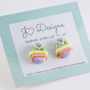 K&ouml;nnte beinhalten: Ein Paar handgefertigte Ohrringe in Form von Ostereiern in Lila und Gr&uuml;n mit gelben und orangen Akzenten. Die Ohrringe werden auf einer Karte mit dem Text "J Designs Handmade pretties just for you!" und einer Website-Adresse pr&auml;sentiert.