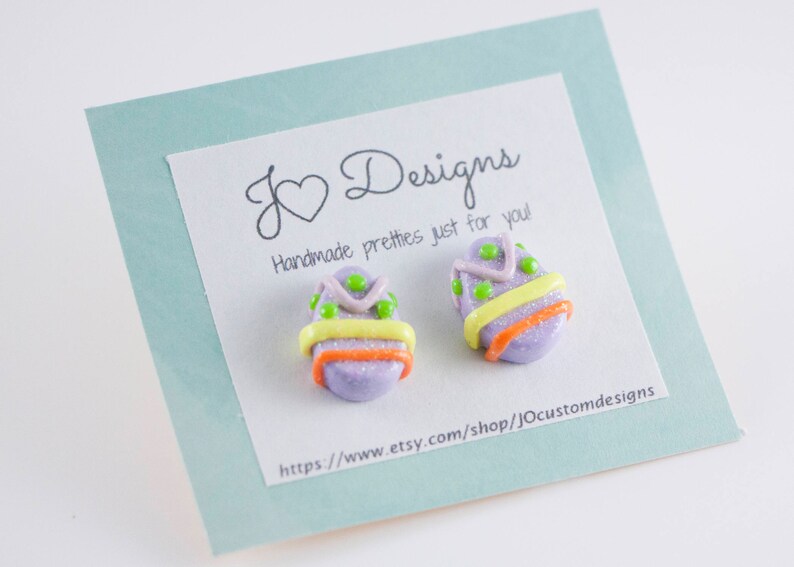 K&ouml;nnte beinhalten: Ein Paar handgefertigte Ohrringe in Form von Ostereiern in Lila und Gr&uuml;n mit gelben und orangen Akzenten. Die Ohrringe werden auf einer Karte mit dem Text "J Designs Handmade pretties just for you!" und einer Website-Adresse pr&auml;sentiert.