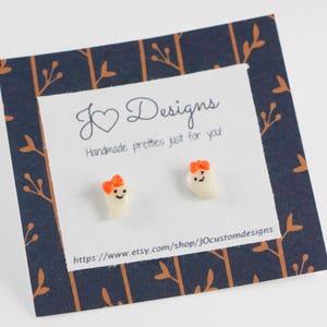 May include: A pair of white ghost earrings with orange bows. The earrings are on a card with the text "J Designs Handmade pretties just for you!" and a website address.