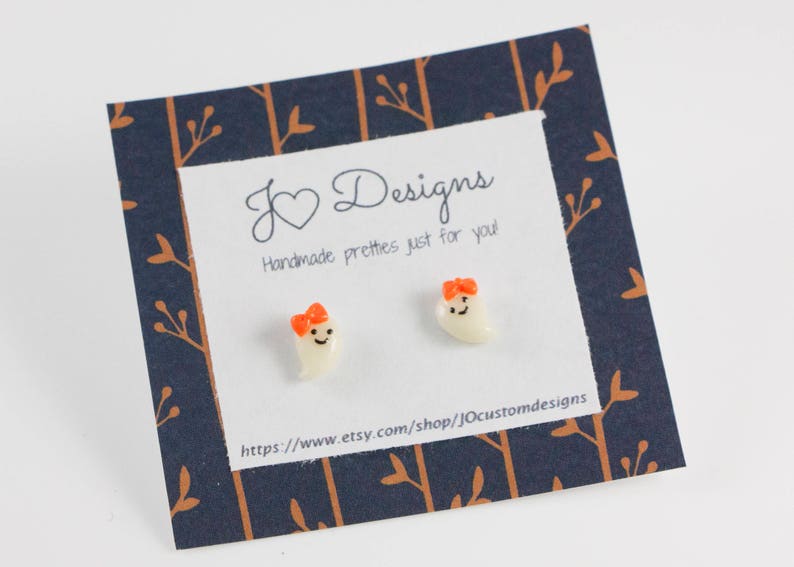 May include: A pair of white ghost earrings with orange bows. The earrings are on a card with the text "J Designs Handmade pretties just for you!" and a website address.