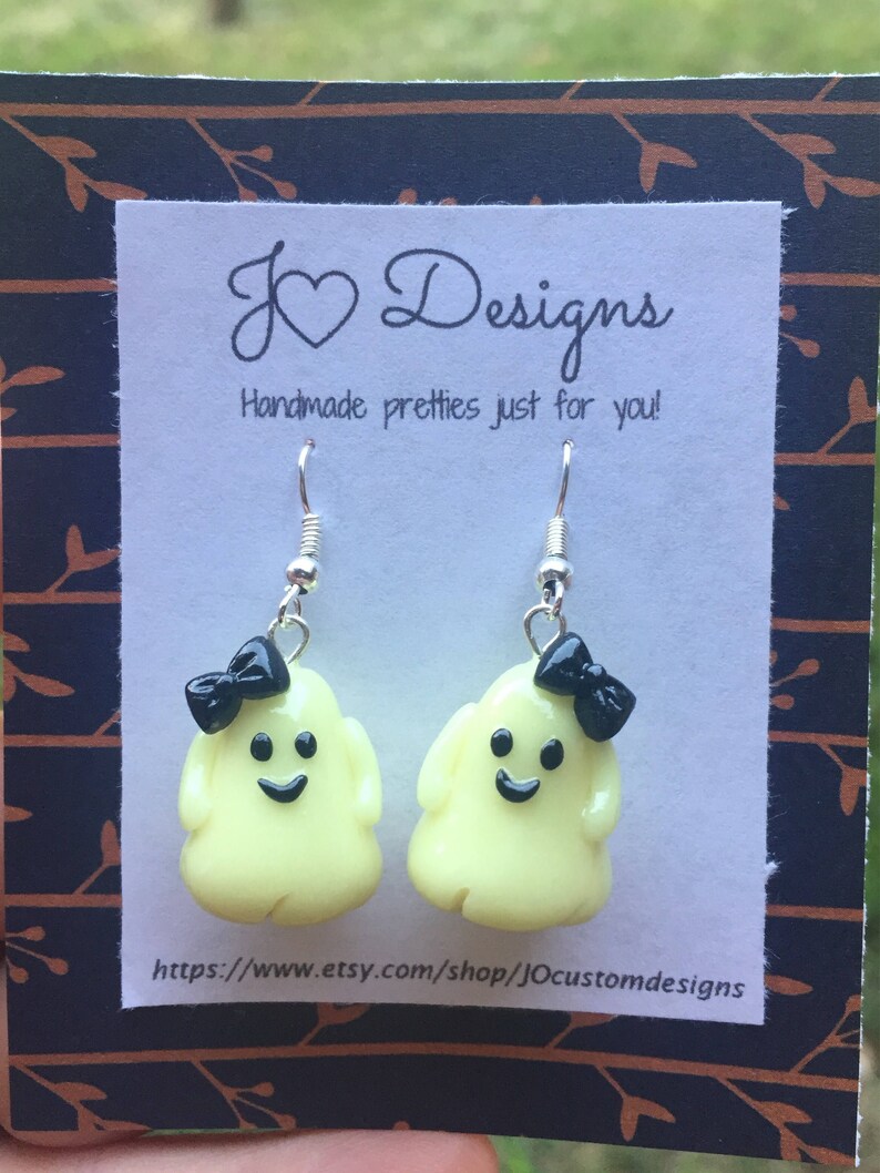 May include: A pair of yellow ghost earrings with black bows and a happy face. The earrings are on a white card with the text "J &hearts; Designs Handmade pretties just for you!" and a website address.