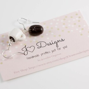 May include: A pair of earrings featuring a white ceramic coffee cup and a brown chocolate-colored donut. The earrings are on a pink background with gold polka dots. The text "J Designs" is written in black cursive script. The text "Handmade pretties just for you!" is written in black. The text "by Jennifer Orgeron" is written in black. The text "Etsy Shop: https://www.etsy.com/shop/JOcustomdesigns" is written in black.